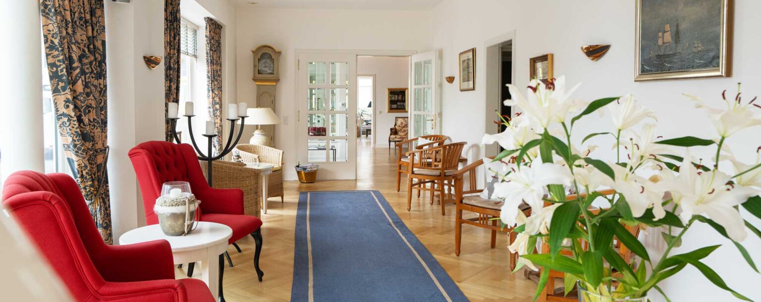 Empfangsraum mit blauem Läuferteppich, roten Polstersesseln, eleganten Holzstühlen, Lilien auf einem Tisch und einer Glastür